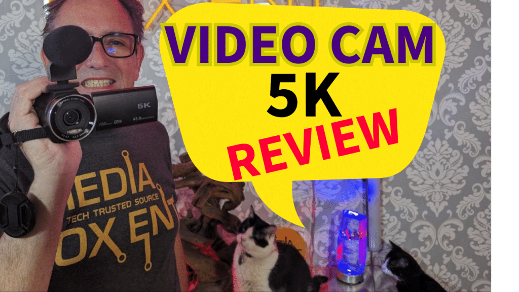 A smiling man holding a 5K video camera with a microphone attachment, standing in front of a patterned wallpaper. A speech bubble graphic with text "VIDEO CAM 5K REVIEW" is prominent. A cat and tech-themed decor are also visible.