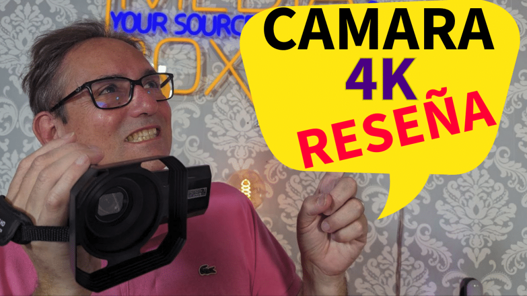 Hombre con gafas sonriendo y sosteniendo una cámara 4K con un gráfico de globo de diálogo que dice 'CÁMARA 4K RESEÑA' en letras negritas.