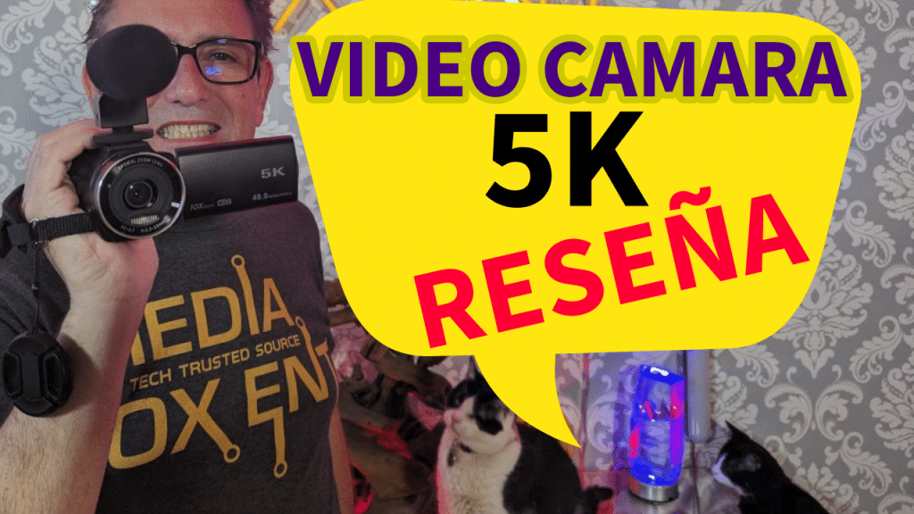Hombre sonriente sosteniendo una cámara de video 5K con texto superpuesto 'VIDEO CAMARA 5K RESEÑA' en una habitación con papel tapiz decorativo y dos gatos.